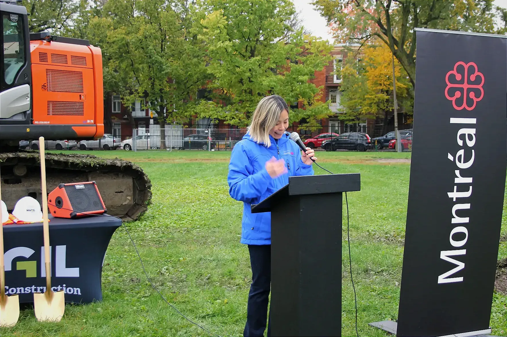La mairesse du Plateau-Mont-Royal, Cathy Wong, parle à un podium à côté d'une bannière de la Ville de Montréal, de pelles, de casques blancs et d'une pelle mécanique dans le parc Baldwin.