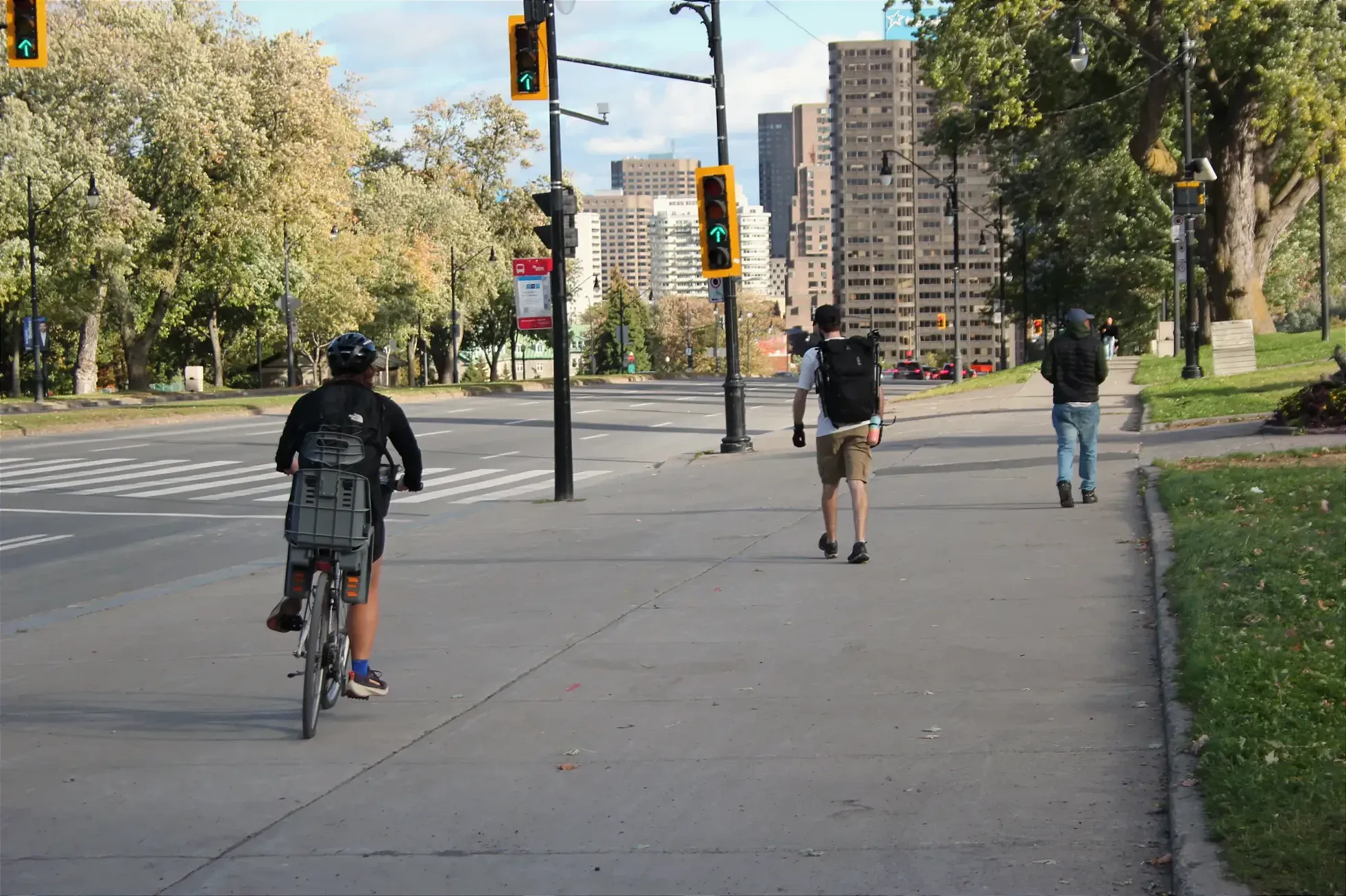 Une cycliste avec un siège de bébé s'apprêt à croiser le chemin de deux piétons qui lui font dos, sur le trottoir de l'avenue du Parc, près du monument à George-Étienne Cartier.