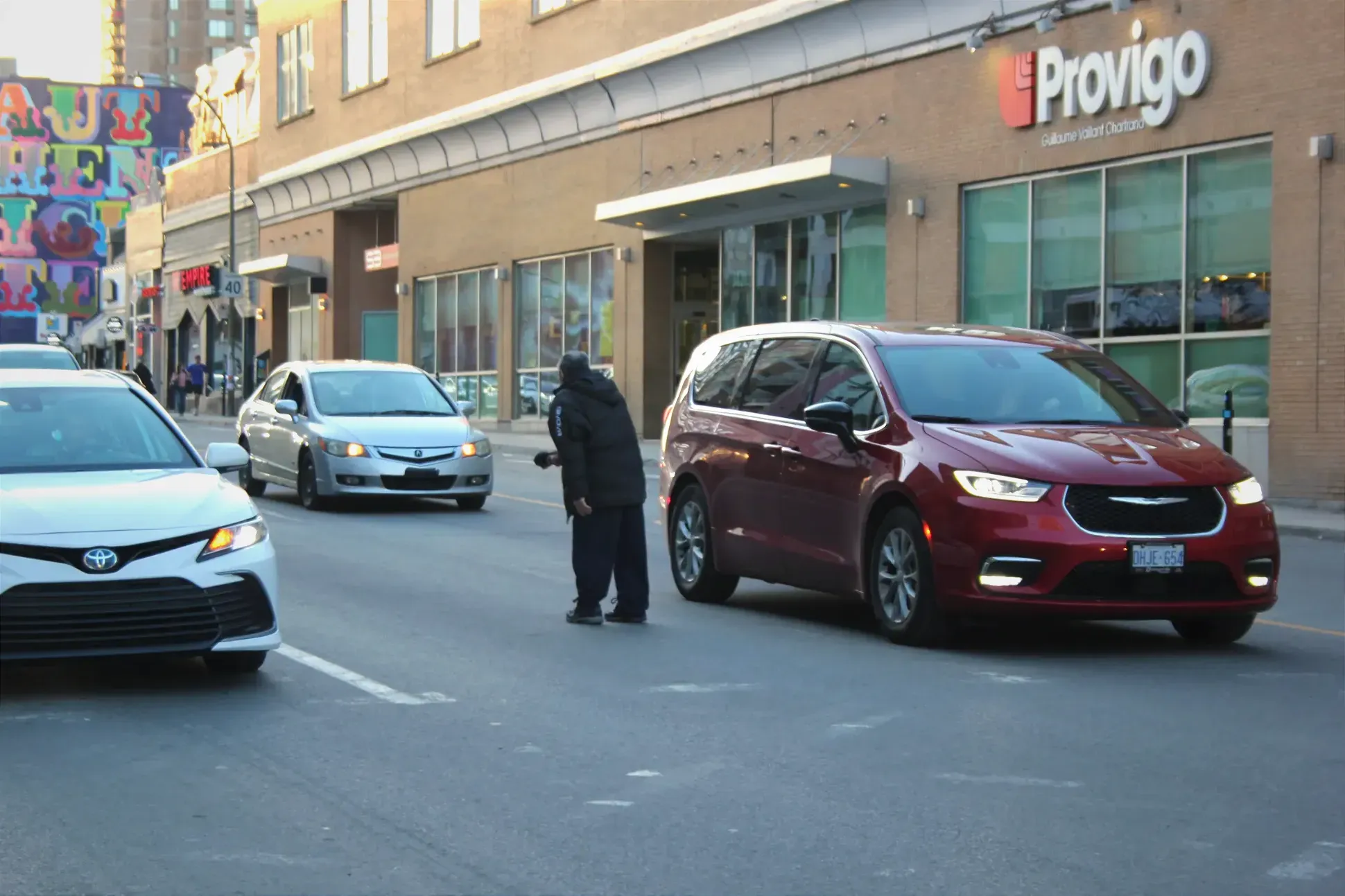 Un homme debout sur une voie, entouré de voitures, fait face au trafic, tendant un chapeau.