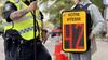 Un agent équipé d'une veste jaune tient un radar d'une main. À côté de lui, sur le trottoir, se trouve une élève à laquelle est accroché un afficheur de vitesse. 