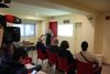 Une photo montre une dizaine de personnes assises dans une petite salle et qui écoutent une femme qui fait une présentation. 