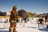 Vêtu d'un costume brun, M. Arnatsiaq tient un tambour noir et rond dans sa main gauche, alors qu'il se promène sur la neige blanche.