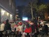 Une foule de dos, dehors le soir, devant une télévision.