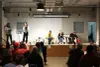 Vue du milieu de la salle presque pleine : une représentante de la table de quartier prend la parole sur scène aux côtés de la modératrice du débat et des trois candidats.