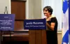 Mme Caserta, debout derrière le podium sur lequel on peut lire le nom de Jennifer Maccarone, semblant émue, tend les mains vers sa poitrine. 