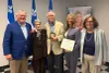 Jacques Chagnon, Phyllis Lambert, Robert Cohen (tenant sa médaille), Jennifer Maccarone (tenant le certificat de M. Cohen) et Charlotte Thibault posent devant des drapeaux du Québec.