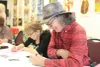 Un homme, crayon à la main, chapeau sur la tête, sourit en pleine rédaction. On peut voir une autre femme à côté de lui, concentrée sur son propre texte.