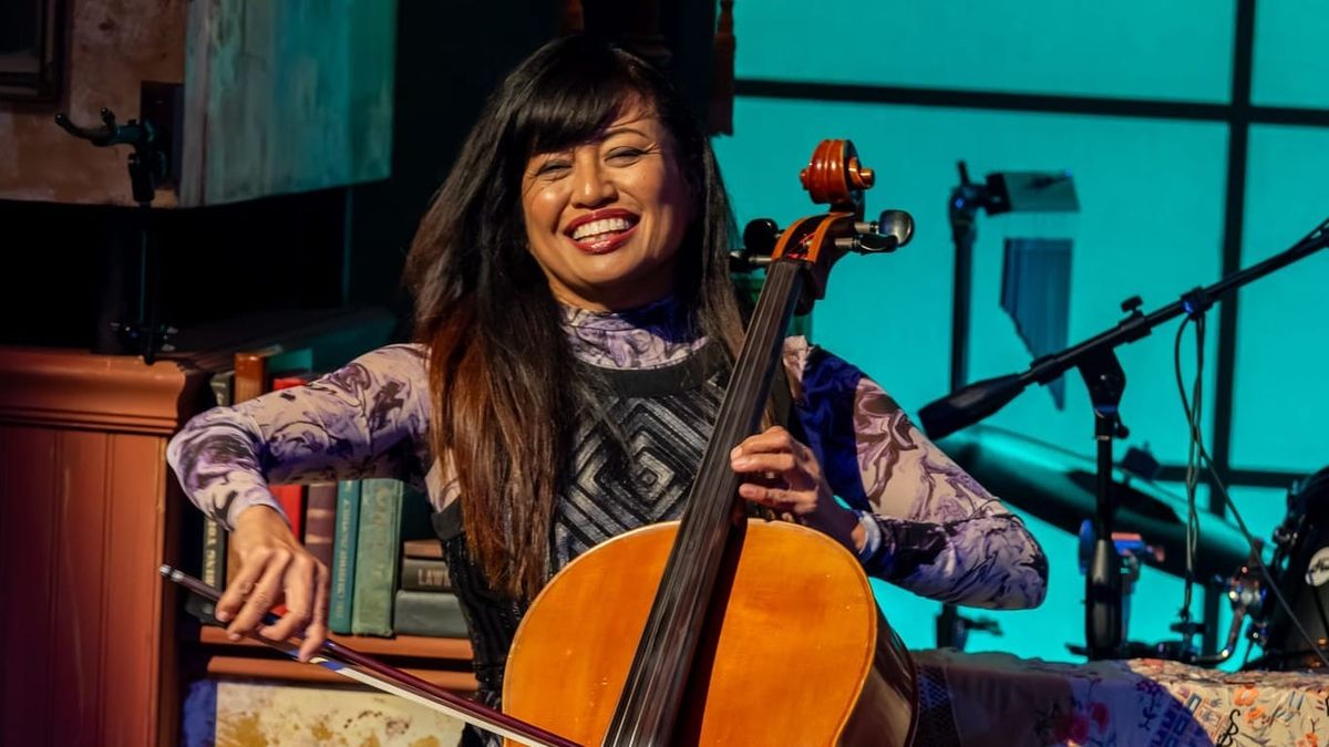 Rose Mallare smiles while playing cello on stage, seated among instruments and vintage décor, with a looping pedal at her feet.