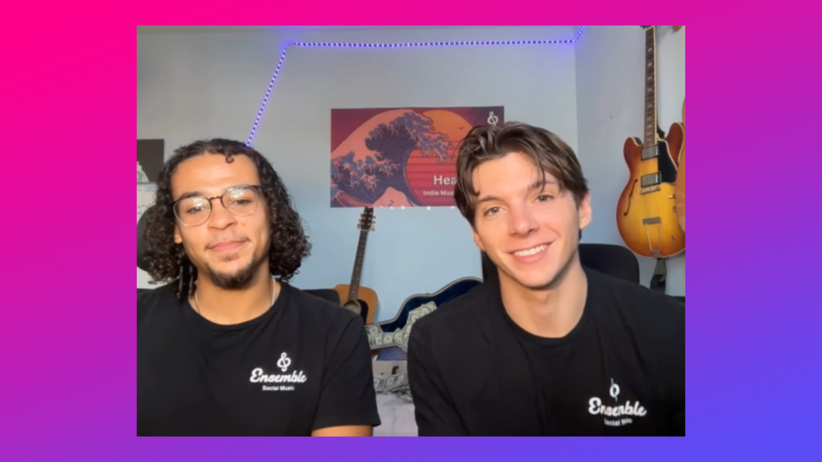 Two young men wearing black Ensemble shirts smile in a music-filled room with guitars on the wall behind them.