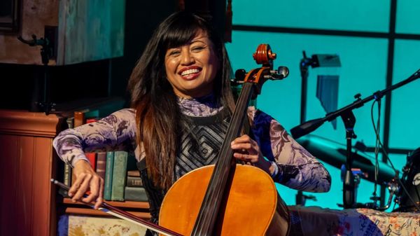 Rose Mallare smiles while playing cello on stage, seated among instruments and vintage décor, with a looping pedal at her feet.
