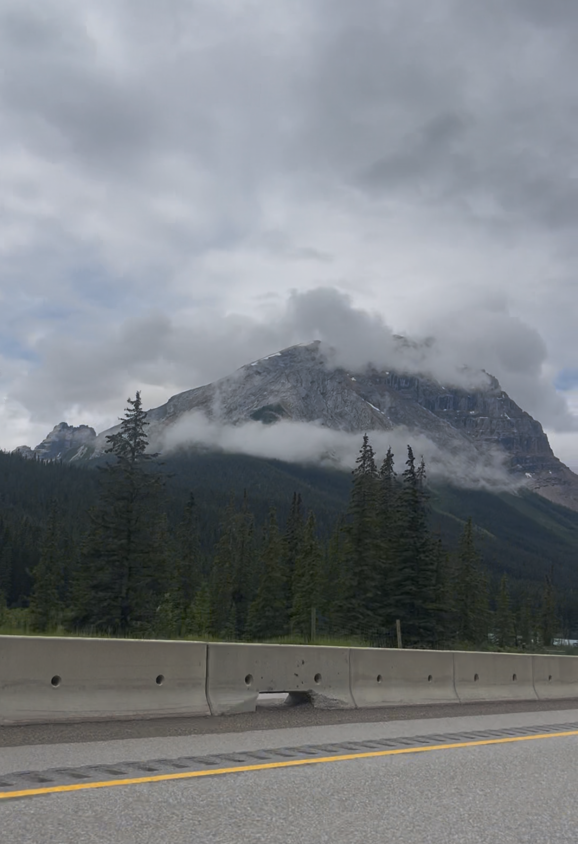 Cloud-wreathed mountain peak rising above the Icefields Parkway, road barrier and yellow centre line in the foreground, Banff National Park