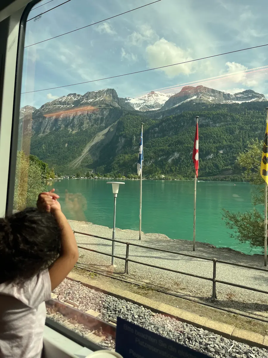 Child at train window looking at turquoise Lake Brienz with snow-capped mountains and Swiss flags, Jungfrau Region Switzerland family train travel