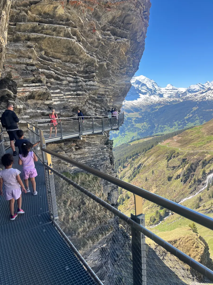 Family on steep mountain staircase path with railing, Grindelwald First Cliff Walk, snow-capped Eiger behind, Jungfrau region