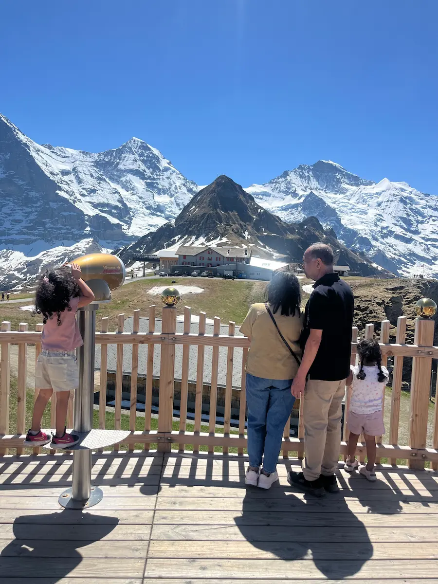Family backs to camera at fenced mountain viewpoint with Eiger Mönch and Jungfrau peaks behind, Männlichen Jungfrau region