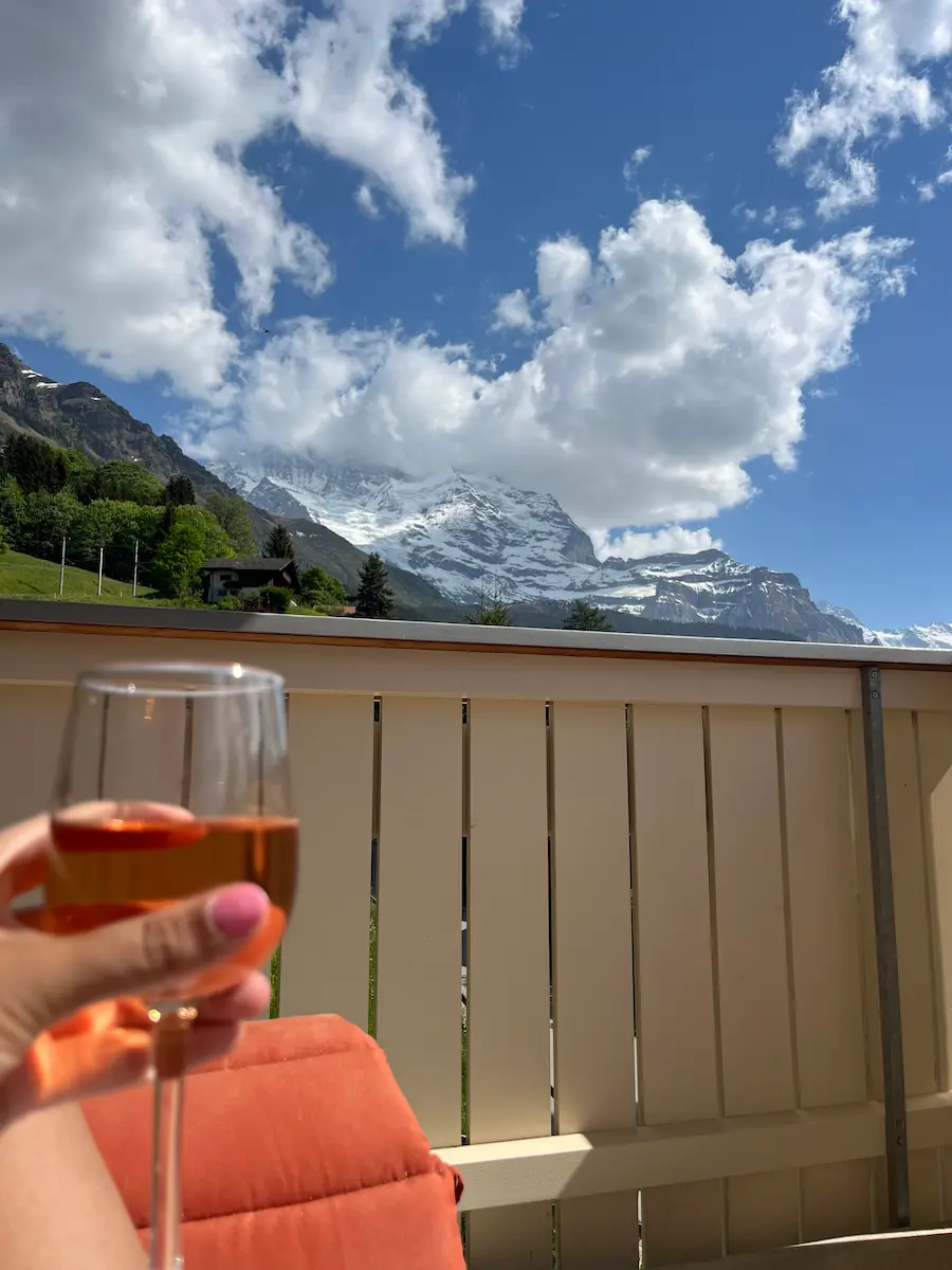 Glass of rosé wine on Wengen apartment balcony panning to snow-capped Swiss Alps evening light, family accommodation Jungfrau region Switzerland