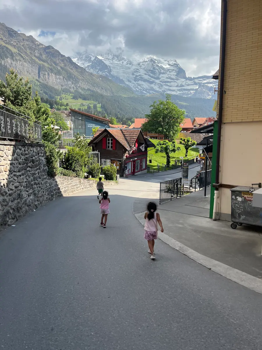 Children walking away backs to camera in Wengen car-free village street with Swiss Alps behind, family travel Switzerland seniors limited mobility