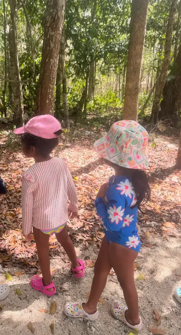 children in sun hats and rash vests walking a jungle path, backs to camera, dappled light through the trees.