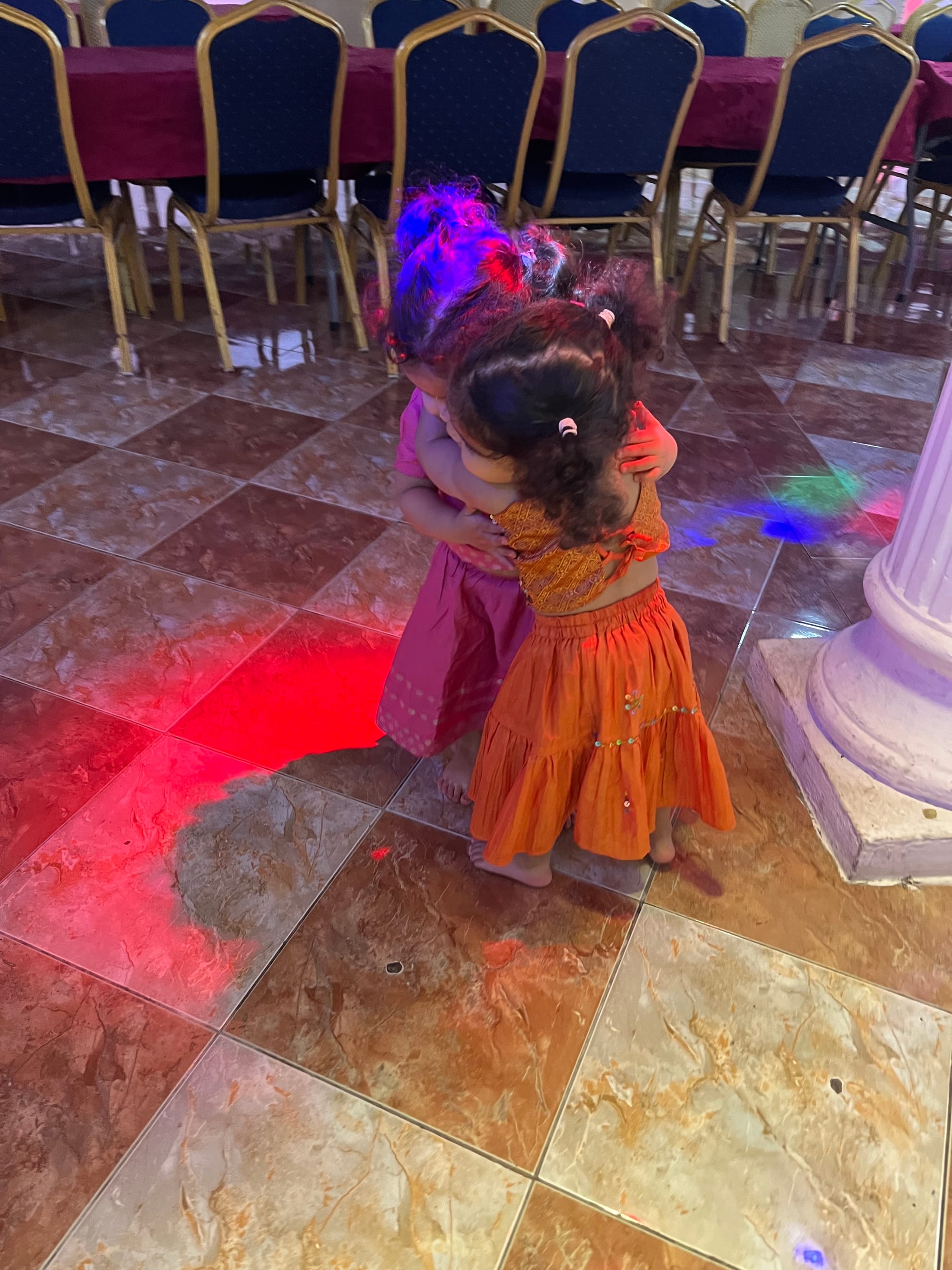 Two young girls in colourful outfits dancing together on a dance floor in Mauritius, backs to camera under coloured lights.