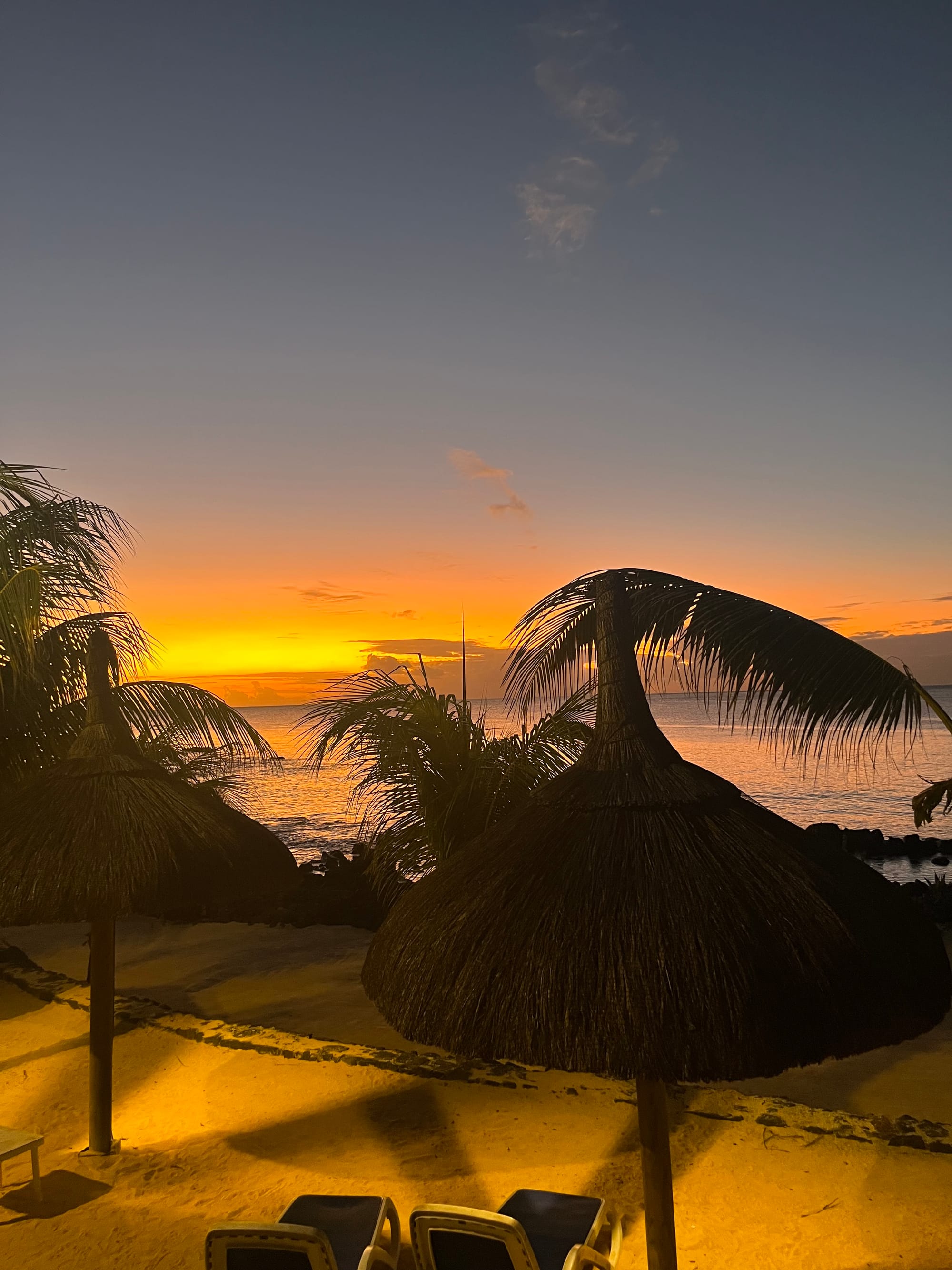 Sunset over the ocean at Flic en Flac, Mauritius, framed by palm trees and thatched beach umbrellas.