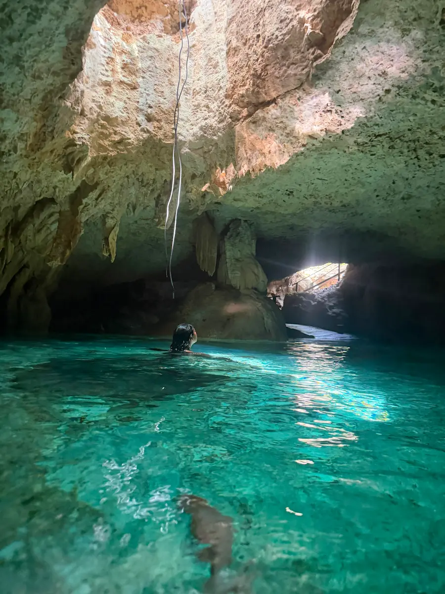 Person swimming through a cave cenote toward a shaft of natural light, stalactites visible overhead, turquoise water below.