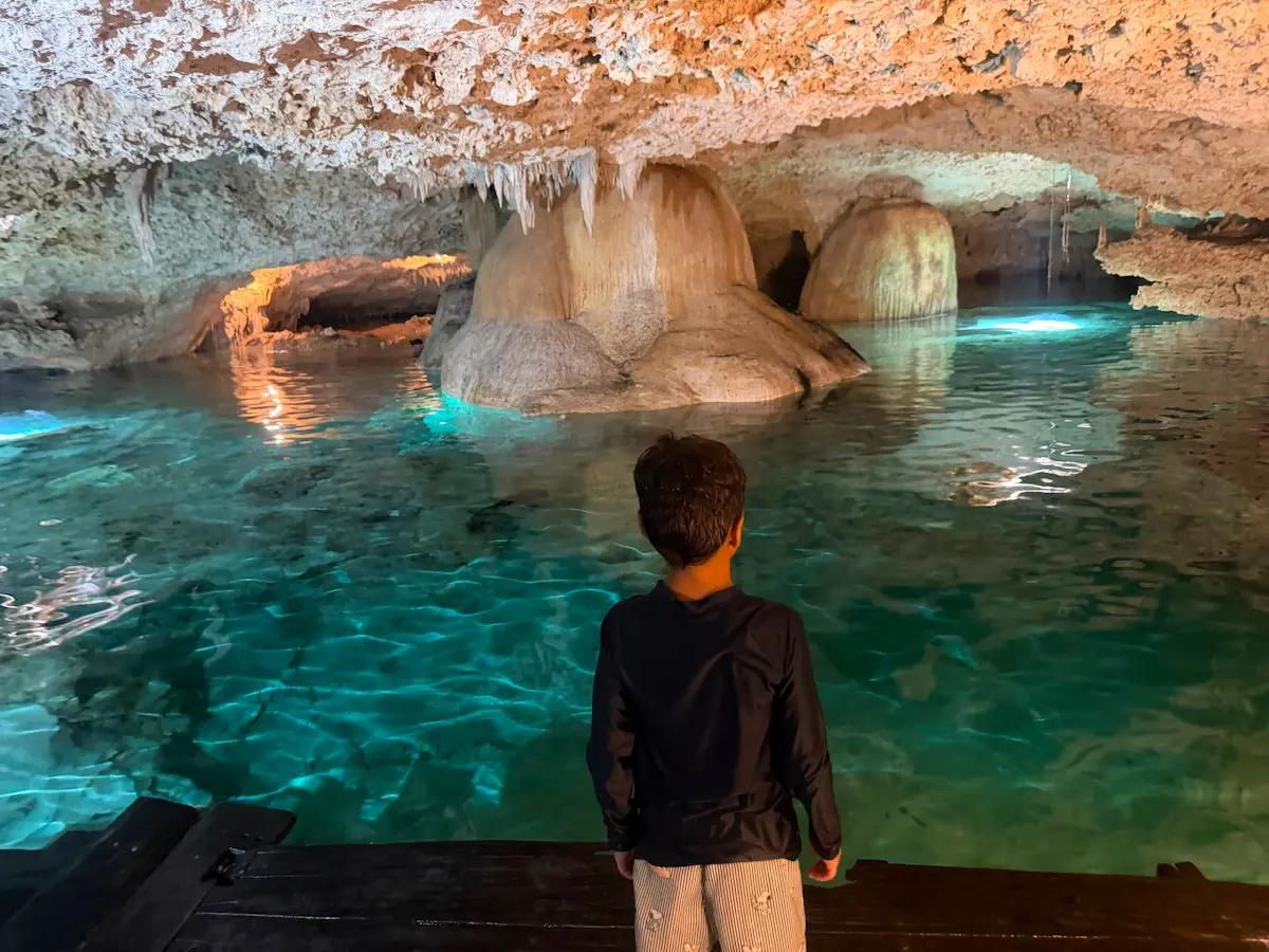 Child standing at the edge of a cave cenote, back to camera, turquoise water and illuminated rock formations ahead.