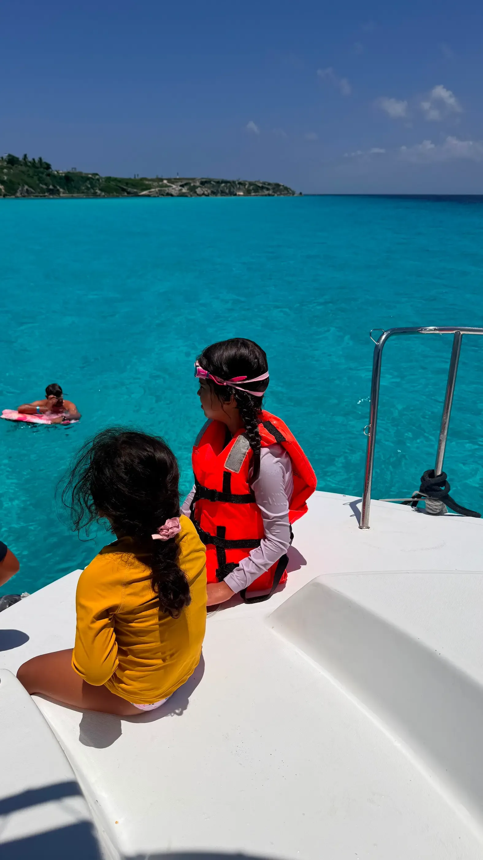 Two children in life vests sitting on the edge of a catamaran, backs to camera, turquoise sea ahead, another swimmer floating nearby.