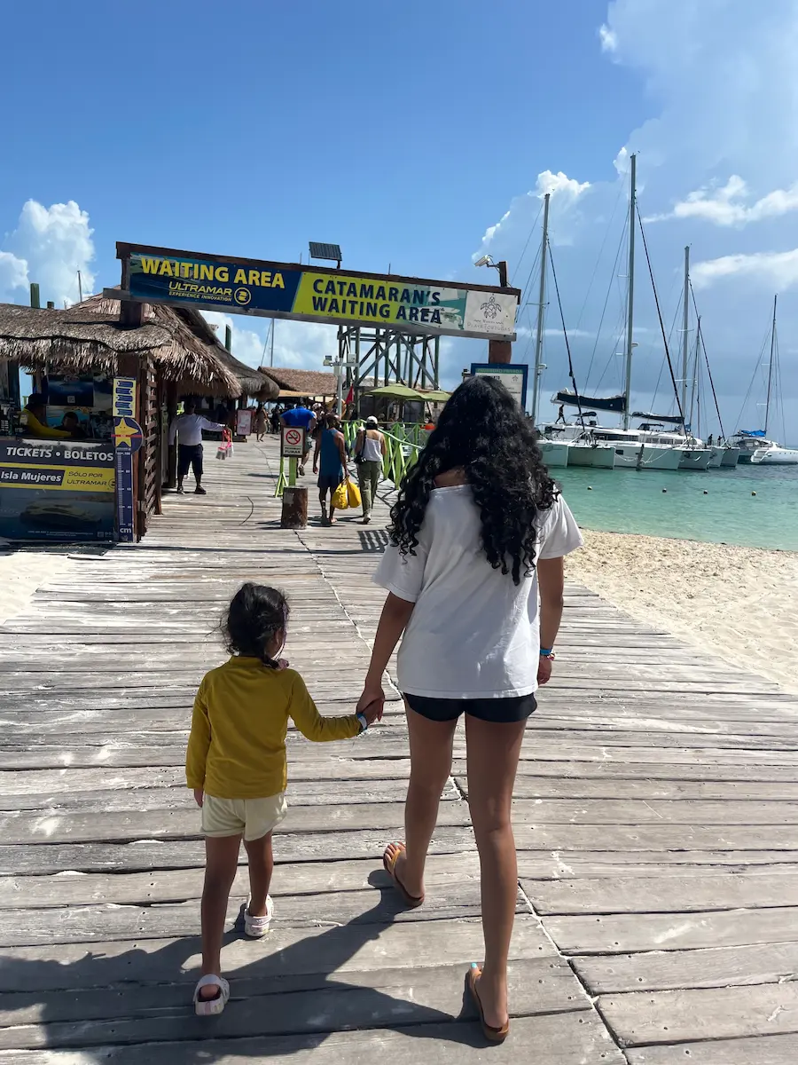 Cousins walking hand in hand along a wooden pier toward the catamaran waiting area, boats visible ahead.