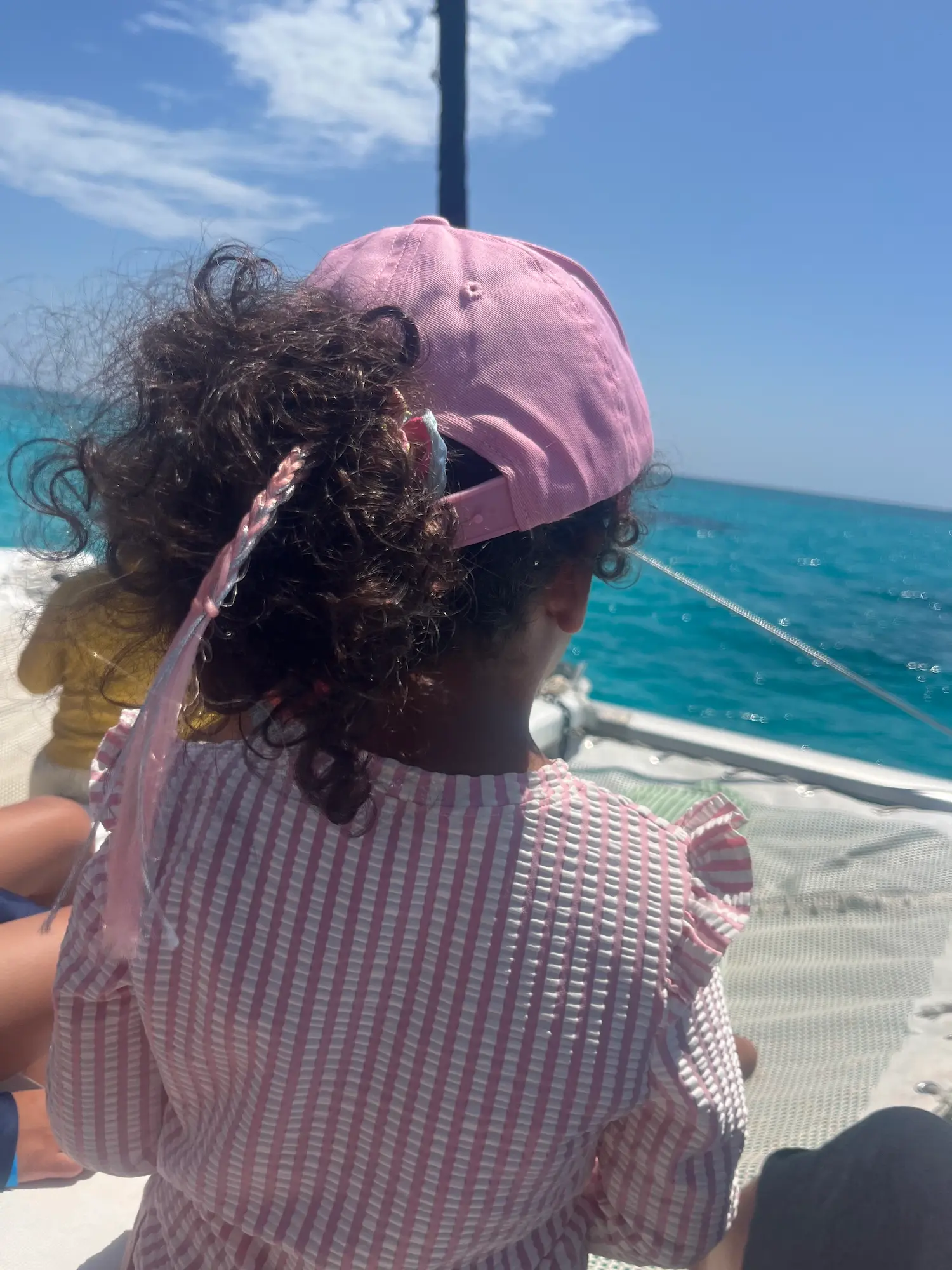 Close view of a child's braided hair and pink cap from behind, seated on a catamaran with the sea beyond.