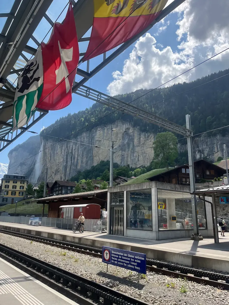 Lauterbrunnen station platform with train tracks, Swiss flags and Staubbach waterfall in background, gateway to Jungfrau Region Switzerland family travel