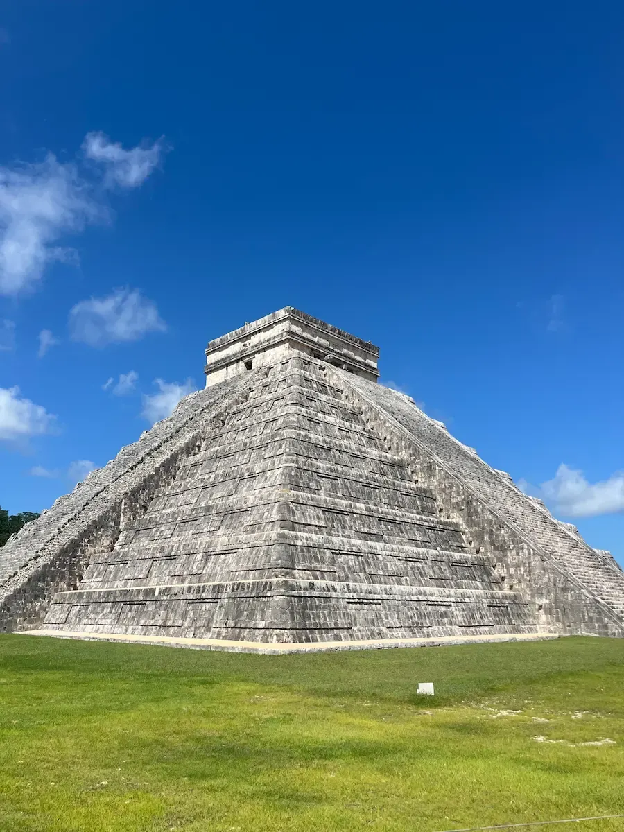 El Castillo pyramid at Chichen Itza under a clear blue sky, full front view with green grass in the foreground.
