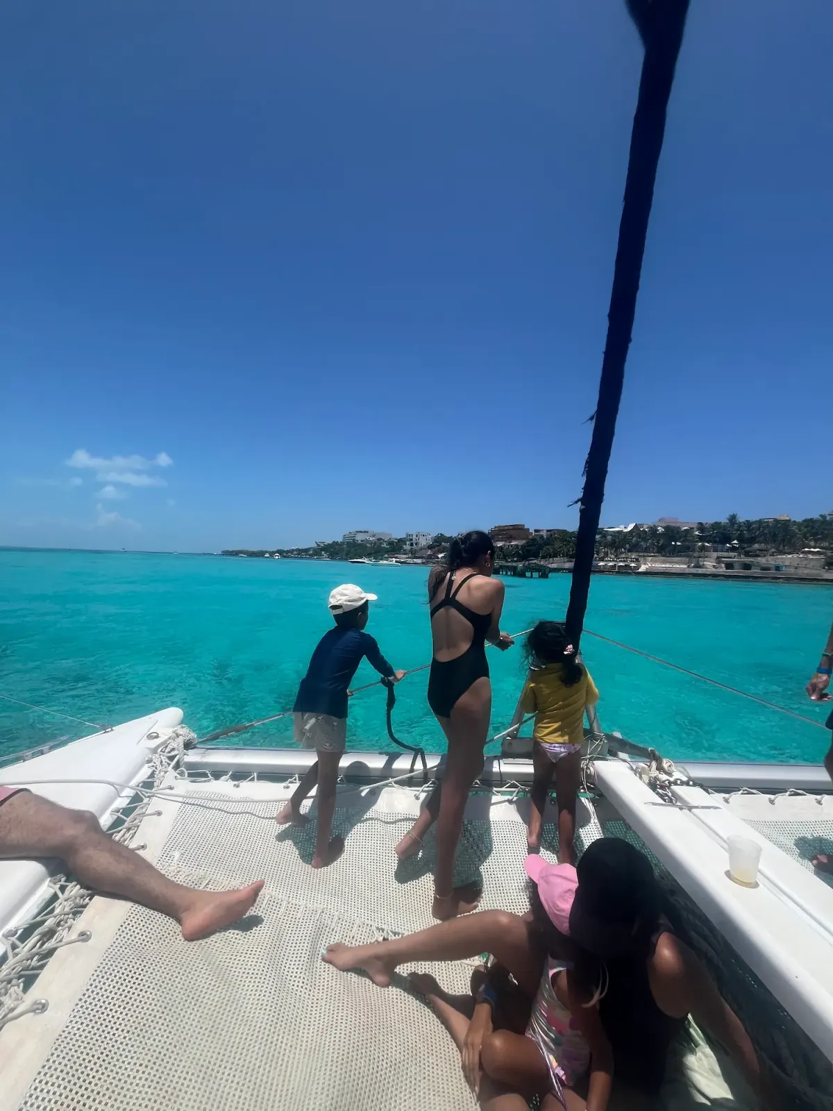 Family standing at the bow rail of a catamaran looking out over turquoise water, backs to camera, resort visible in the distance.