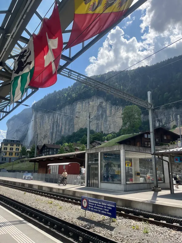 Lauterbrunnen station platform with train tracks, Swiss flags and Staubbach waterfall in background, gateway to Jungfrau Region Switzerland family travel