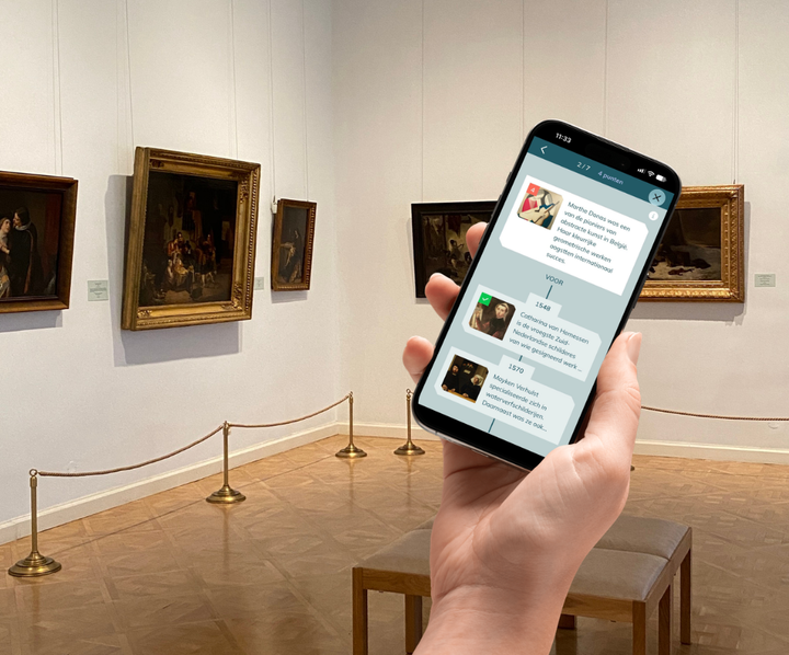 A hand is holding a phone displaying the ErfgoedApp. In the background, we see a museum gallery.