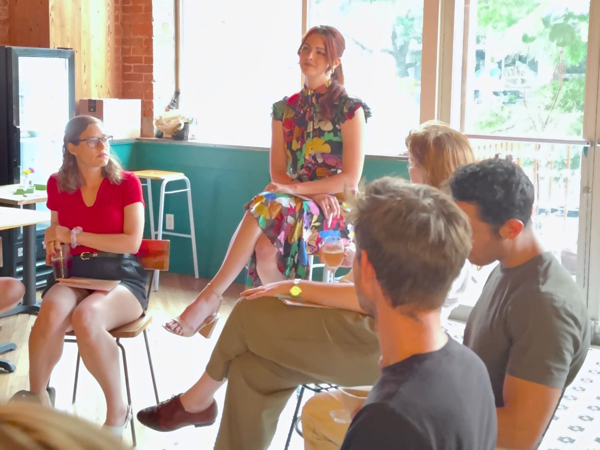 Aubrey sits on a high stool in a colorful dress, speaking to a small group of participants seated in a casual cafe-style space during her Open Loops event in New York City