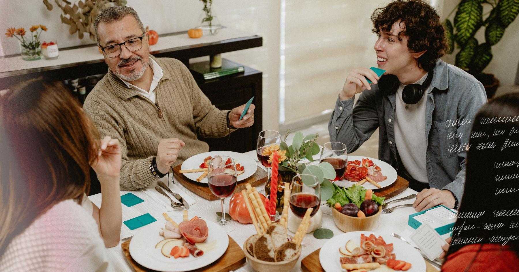 Picture of a family laughing on a thanksgiving table, playing a card game with glasses of wine, candles, a basket of bread and salami plates