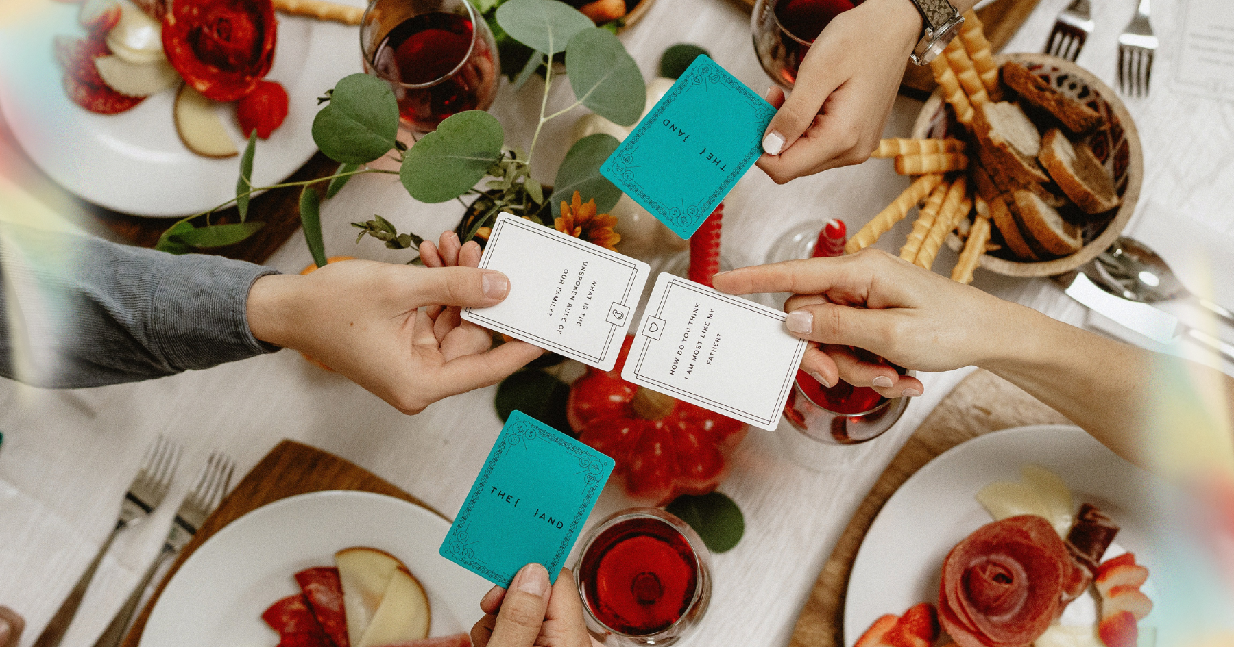 Picture of a card game for families on a thanksgiving table with glasses of wine, hands holding cards, a basket of bread and salami plates