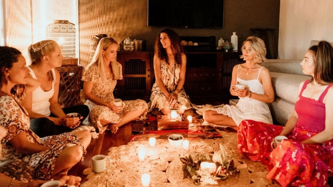A group of six women sitting in a candlelit circle on the floor, talking and holding mugs during an intimate evening gathering.