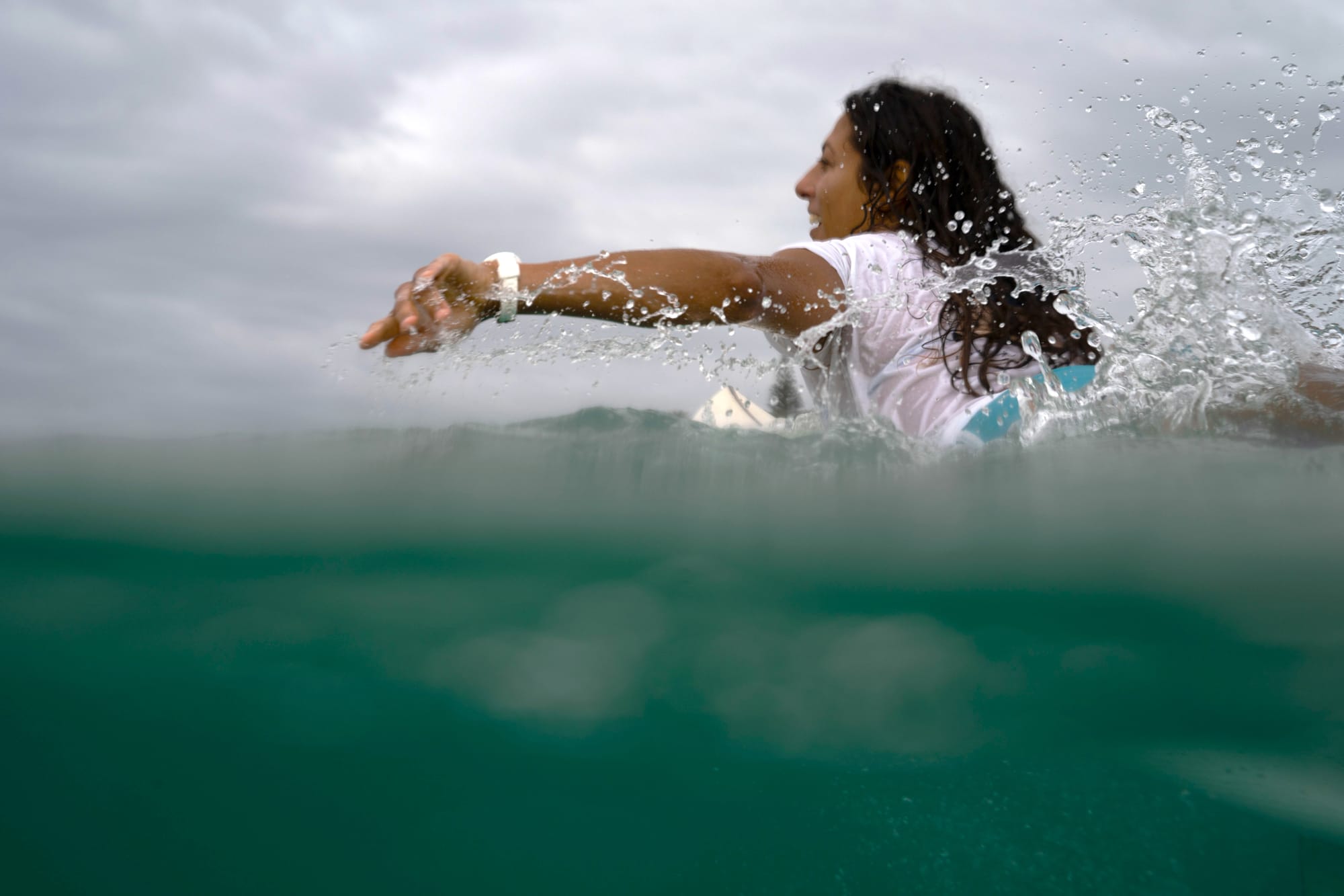 Sonia Houria Essia Rivas paddling on a surfboard in the ocean, mid-motion with water splashing around her.