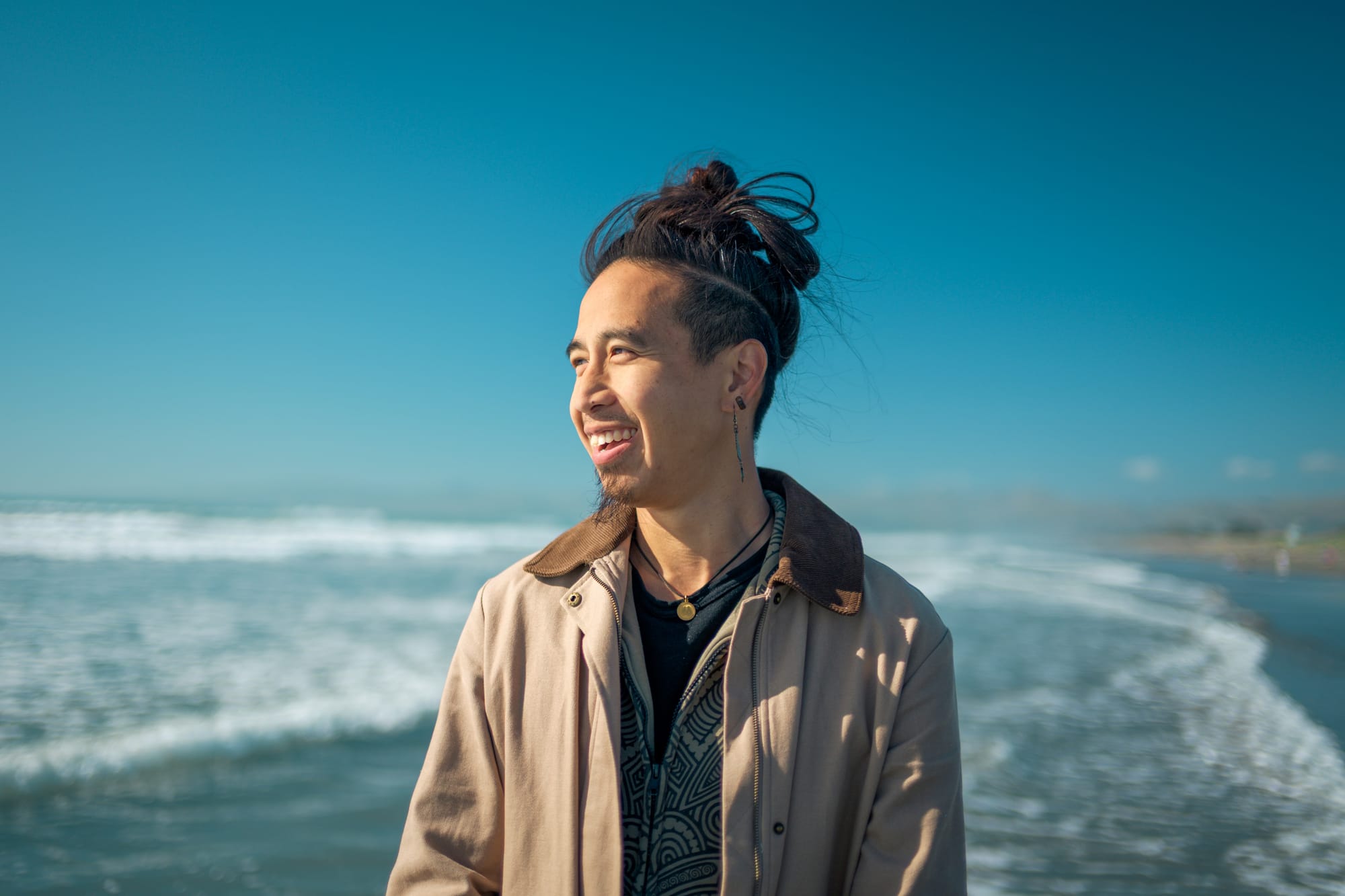 Kai J. Lee (李曉敦) smiling and looking off-camera at the beach, wearing a tan corduroy-collar jacket, with ocean waves in the background