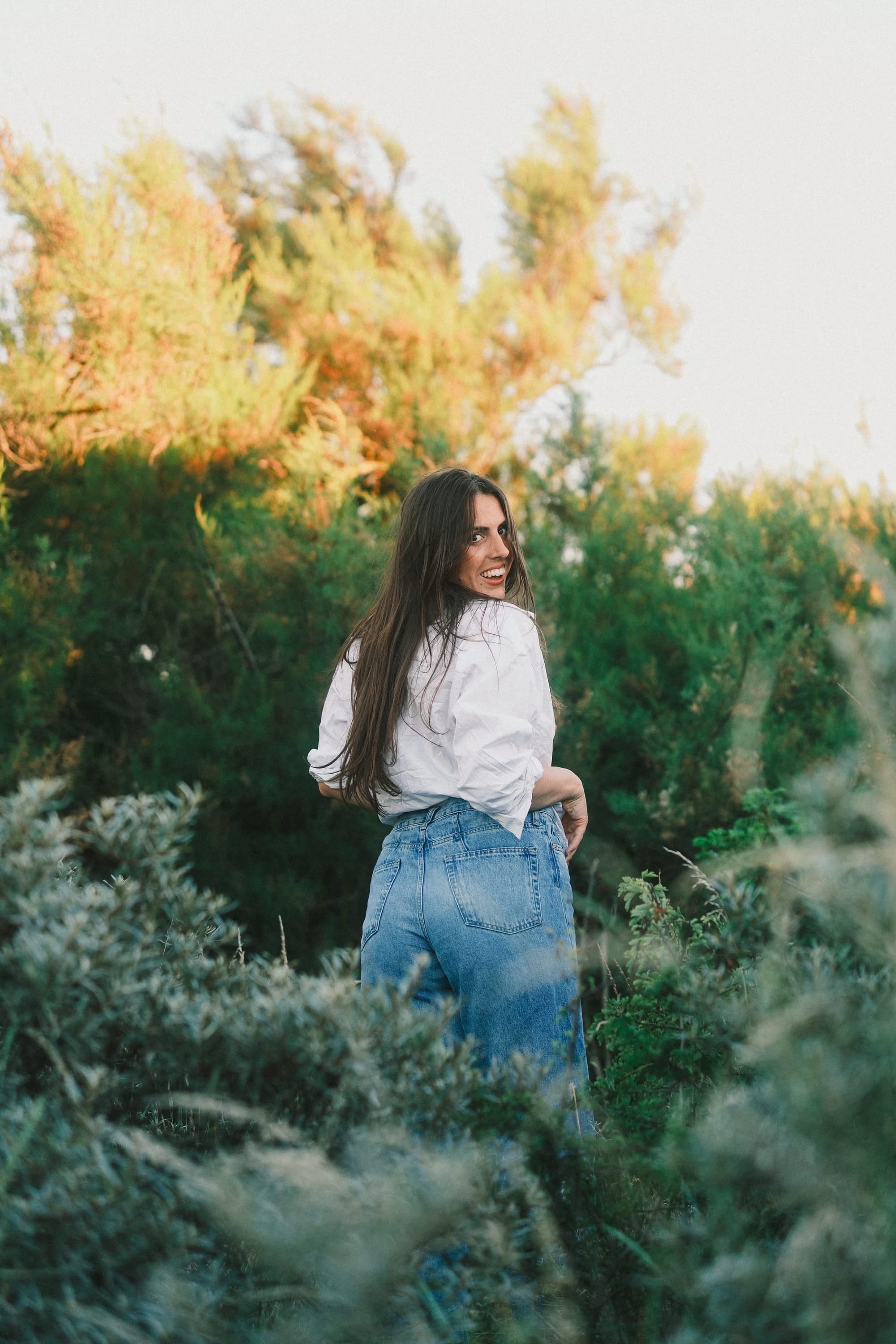Angie Inghelram standing in a field of greenery at golden hour, looking back over her shoulder and smiling, wearing a white blouse and blue jeans.