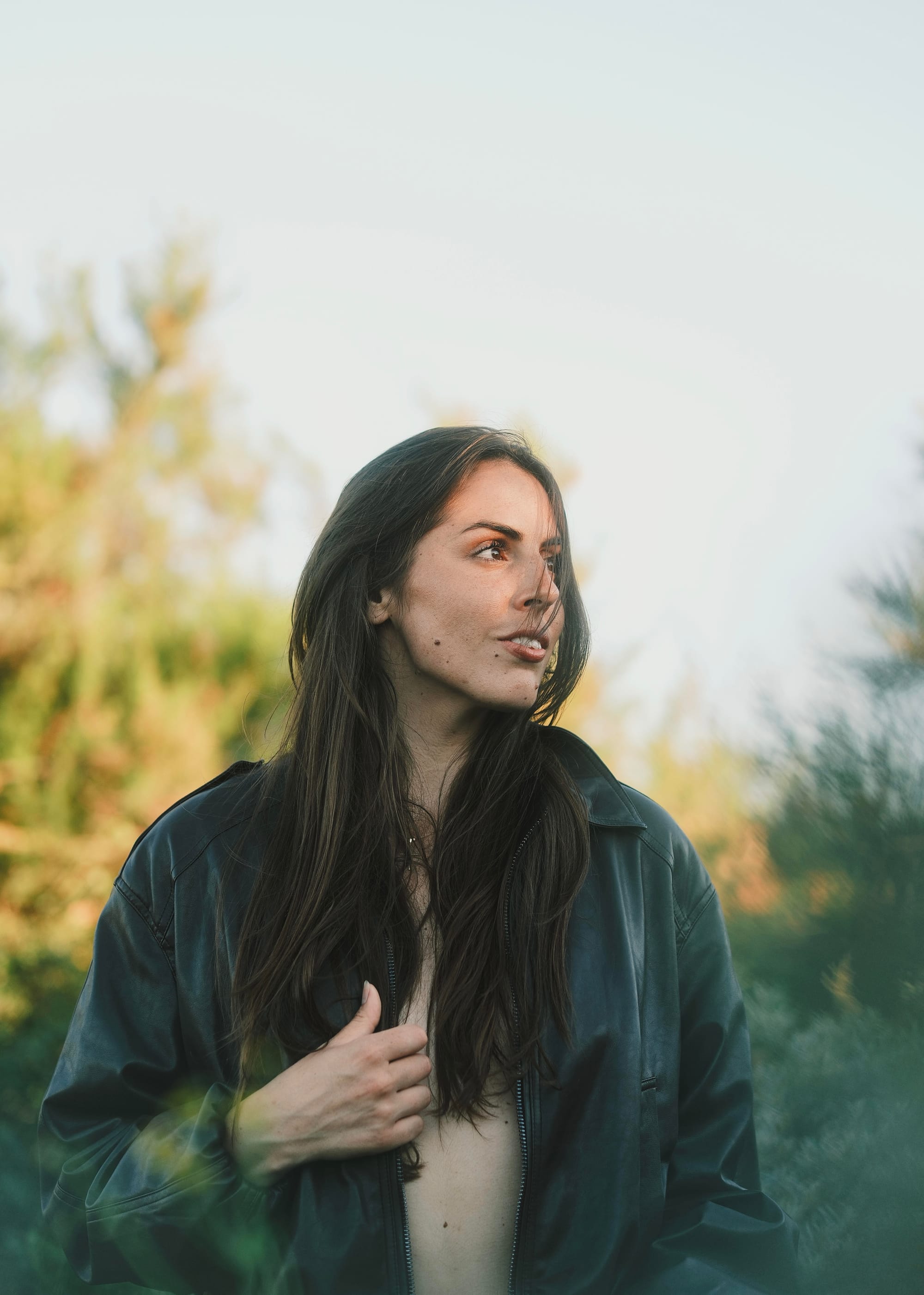 Angie Inghelram standing outdoors at golden hour, looking off to the side, wearing an open black jacket with greenery and trees blurred in the background.