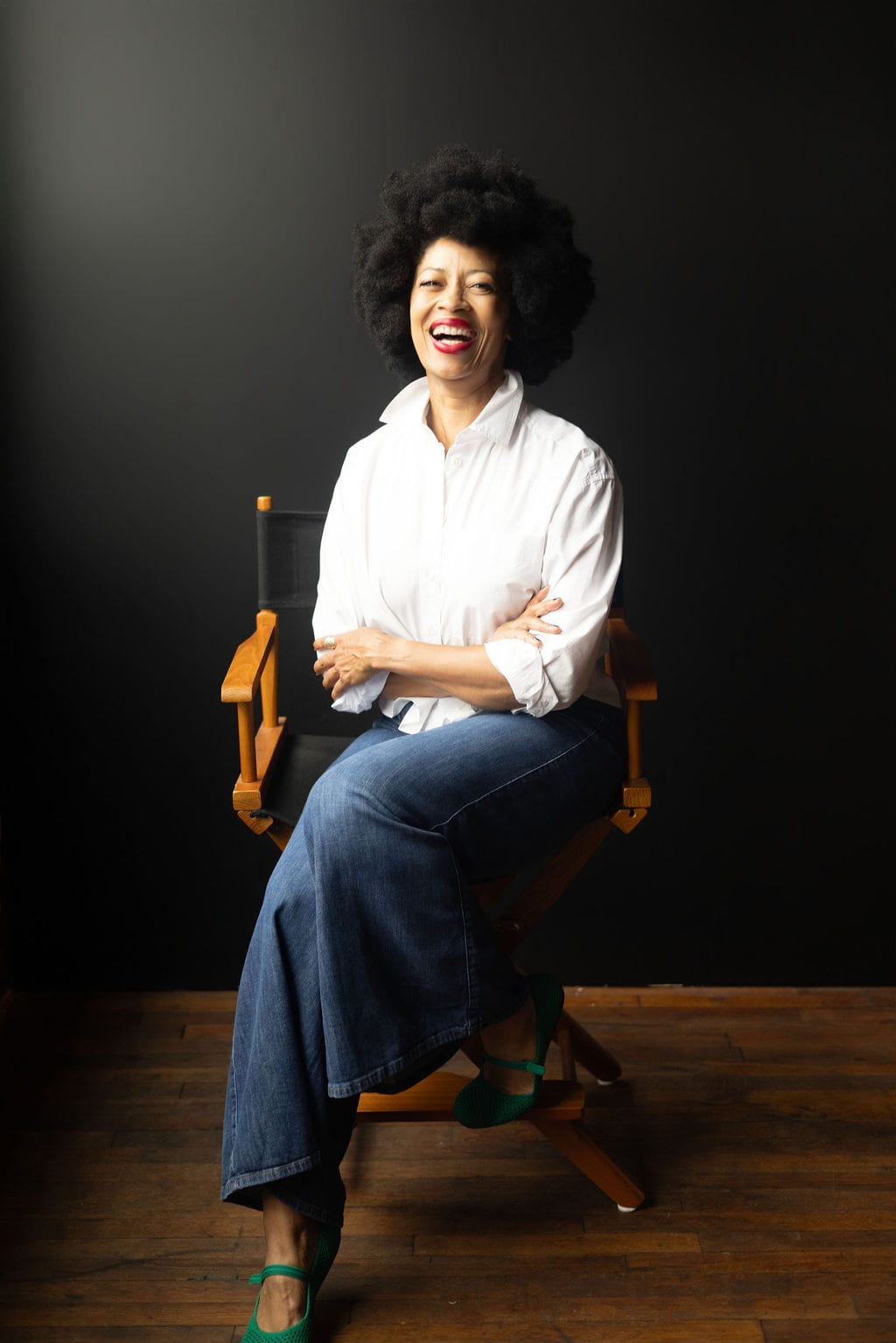 Lenna Davis laughing while seated in a director's chair against a dark backdrop, wearing a white button-down shirt, wide-leg denim, and green flats.