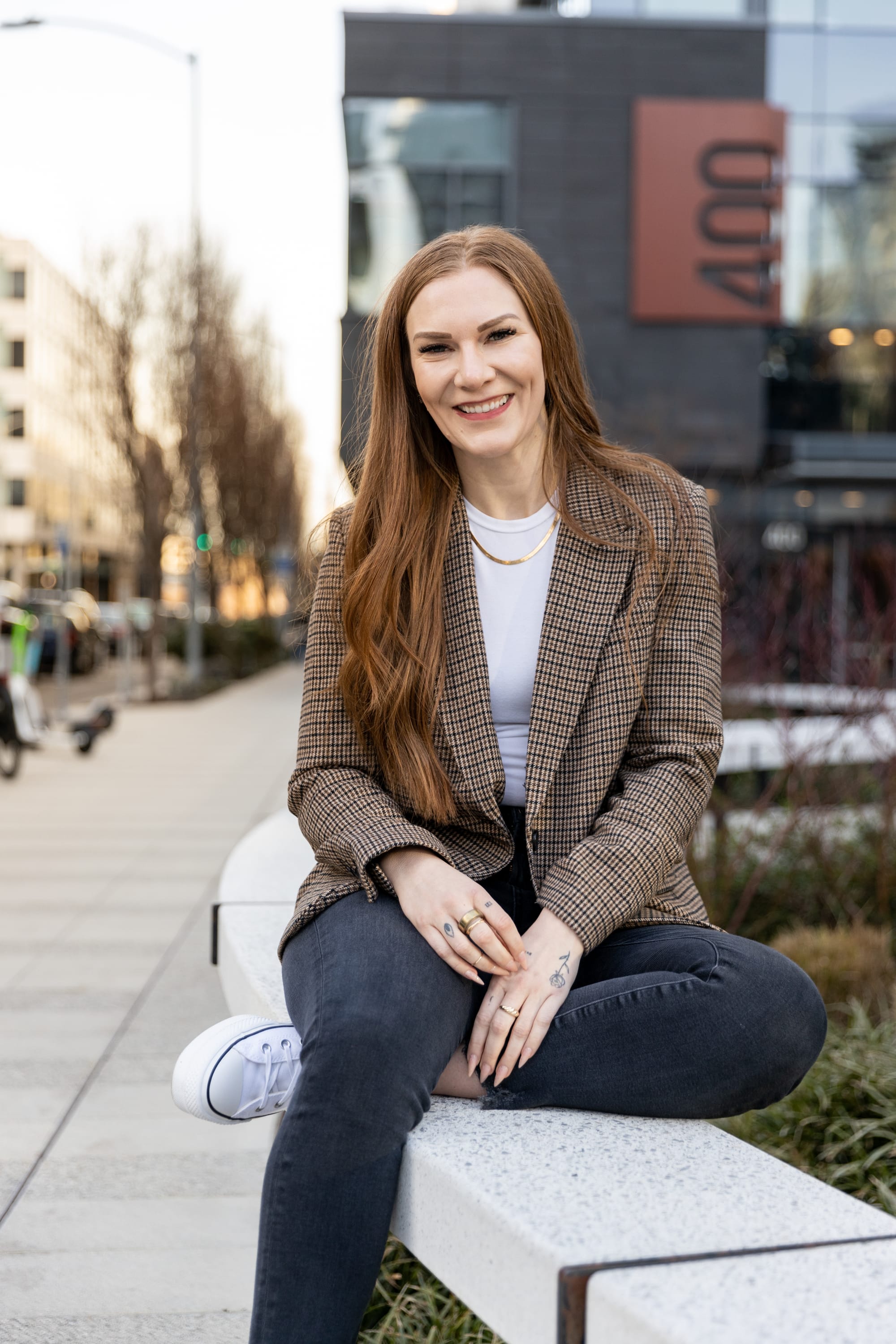 Jennica Lynn sitting cross-legged on a bench in downtown Seattle, smiling in a plaid blazer and jeans.
