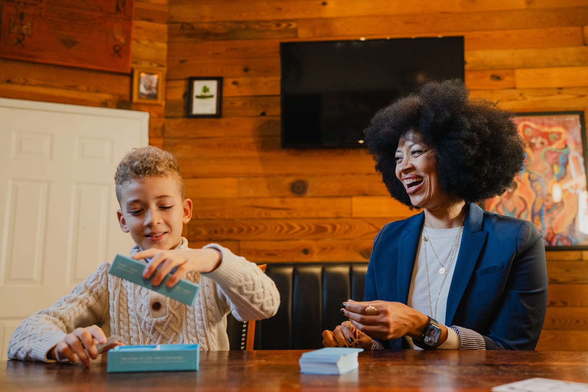 Lenna Davis laughing while sitting at a wooden table with her son Pierre, both holding cards from The Skin Deep's Kids edition of {THE AND}.
