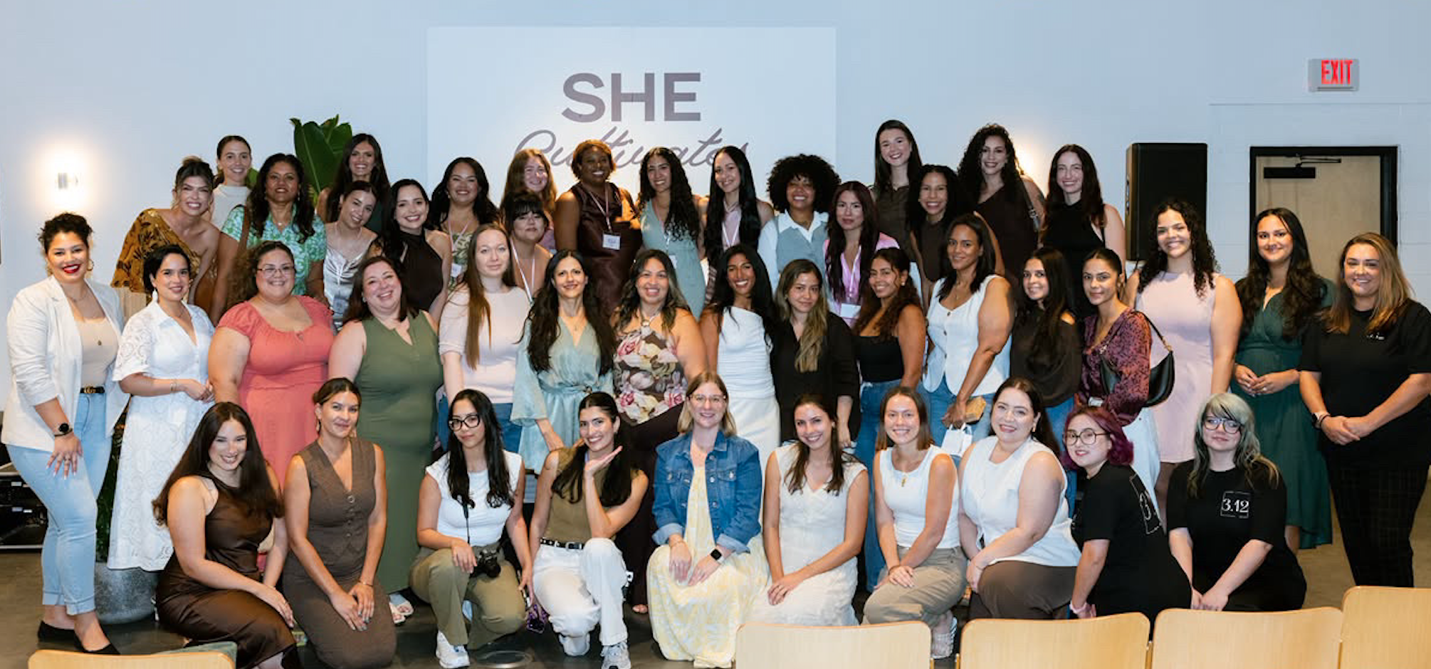 A large group of women posing together at a She Cultivates event, standing and kneeling in rows in front of a screen with the She Cultivates logo.