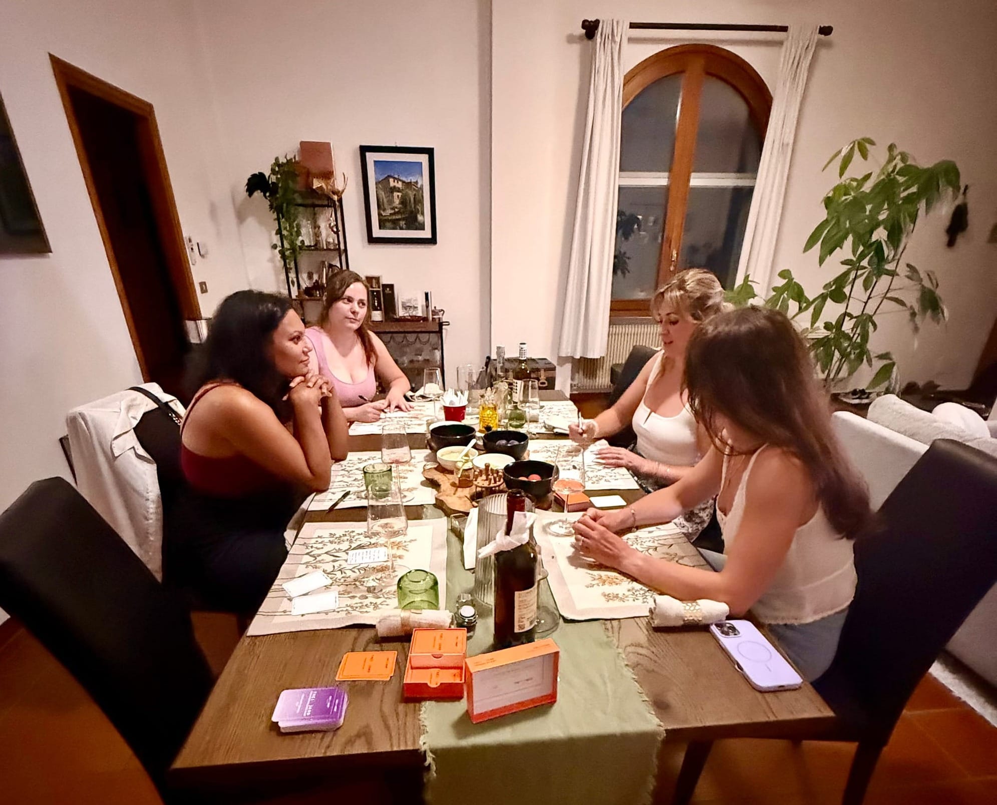 Four women gathered around a dining table at Missy's home in Sacile, Italy, playing {THE AND} card game together over wine and food.