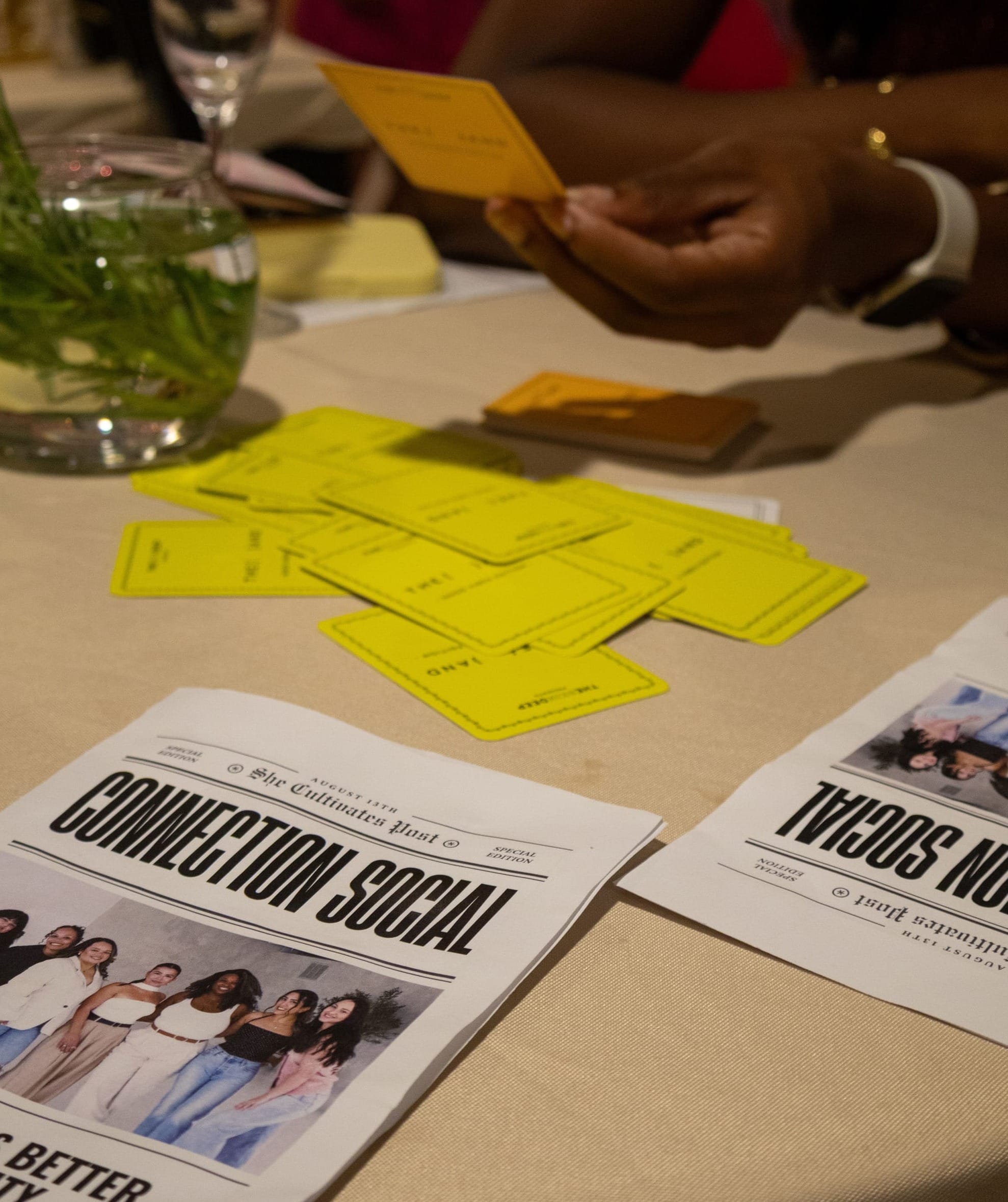 Close-up of a table at Elizabeth's Orlando event with yellow {THE AND} cards spread out, a "Connection Social" printed program, and a guest holding a card.