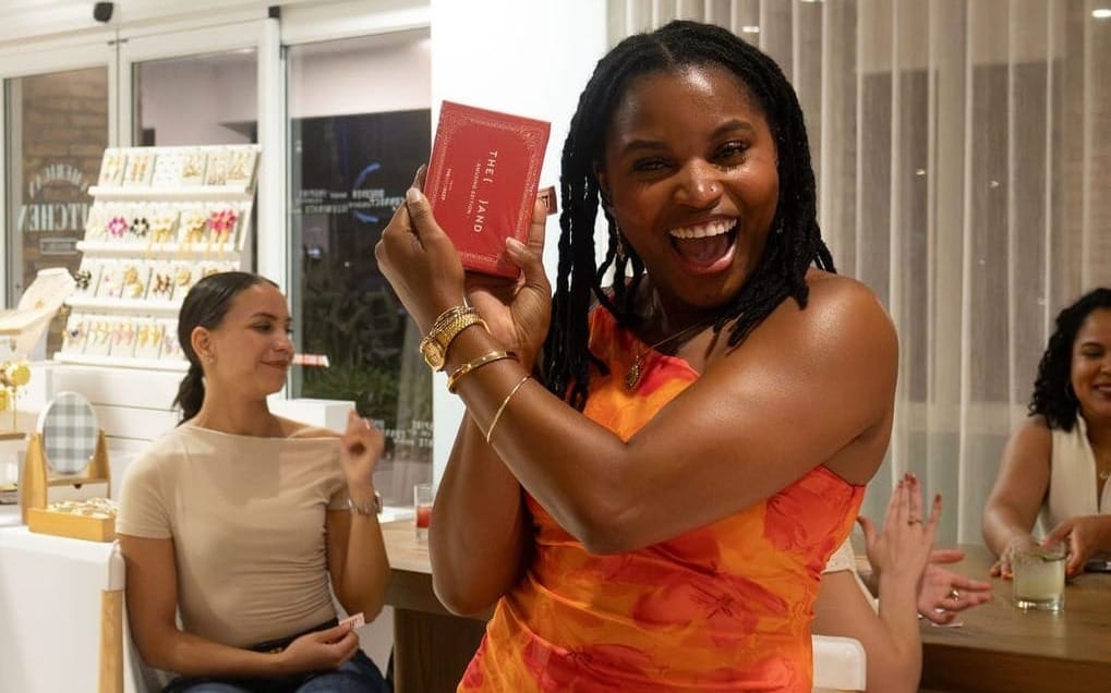 A radiant woman in an orange floral dress joyfully holding up {THE AND}'s Amusing edition at Elizabeth's Orlando gathering, with guests smiling behind her.