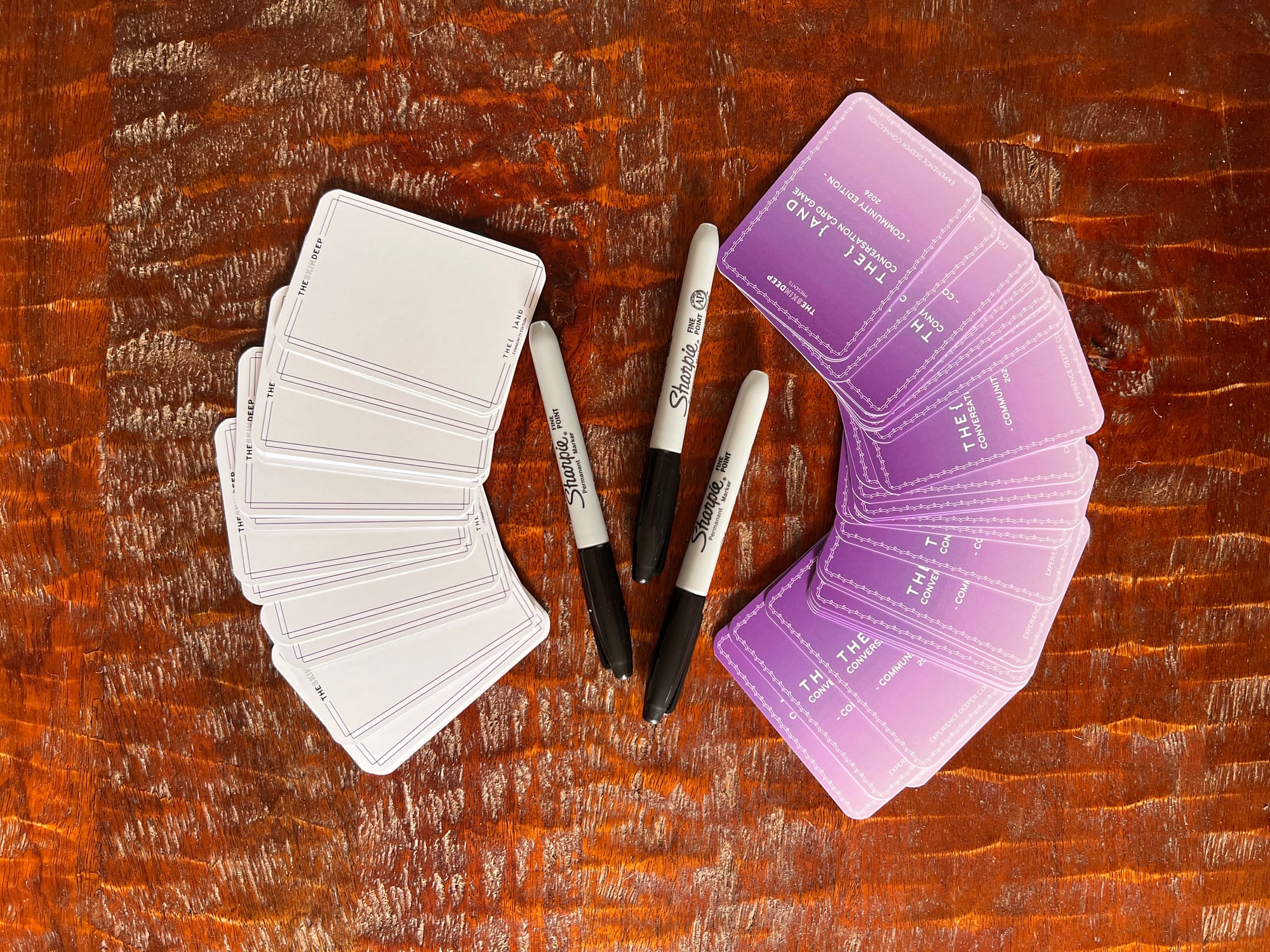 Blank cards and purple 2026 {THE AND} Community Edition cards fanned out on a wooden table alongside Sharpie markers at Liz's Denver gathering.
