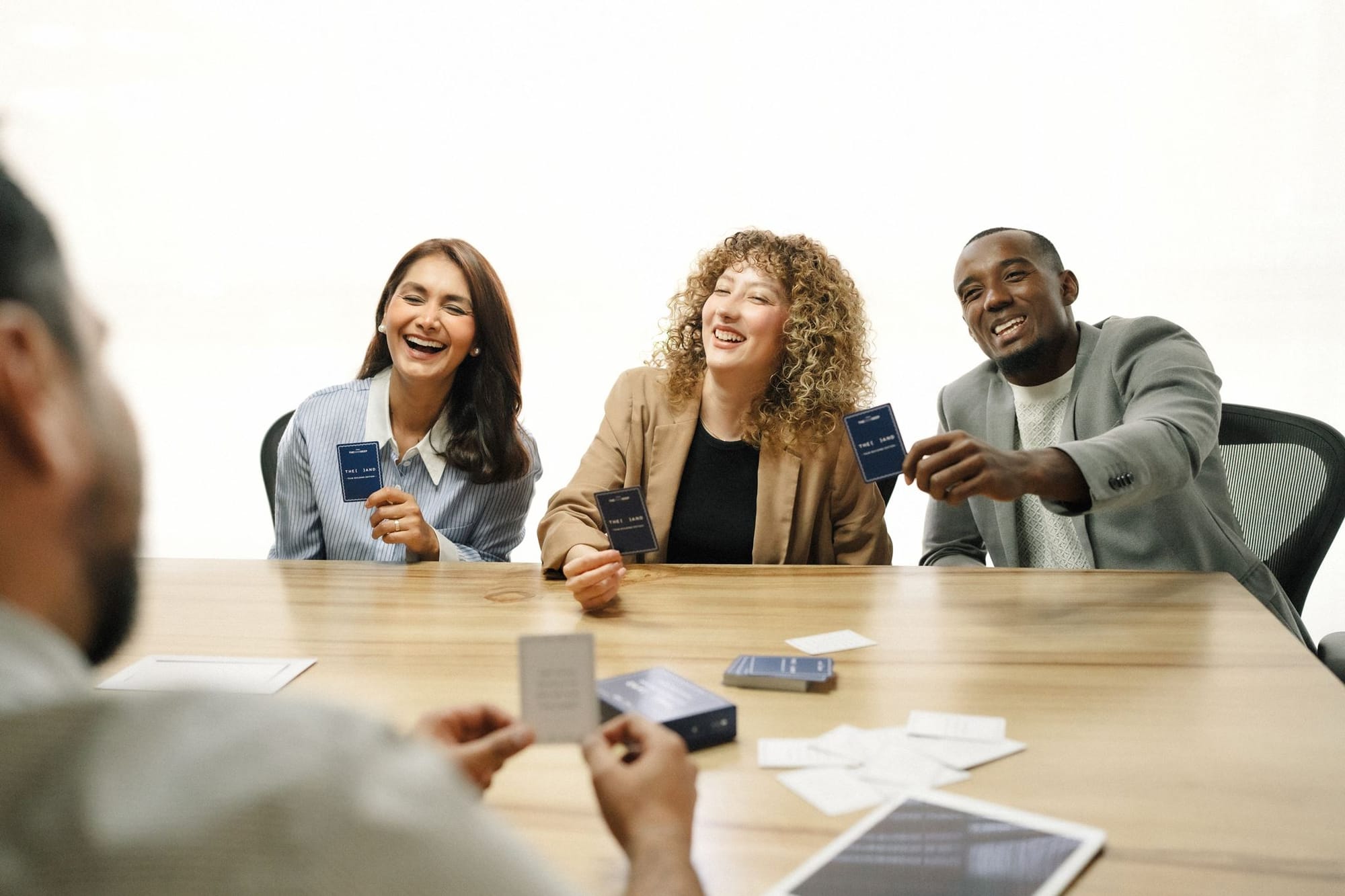 Three coworkers laughing around a conference table, holding cards from {THE AND} Team Building Edition.